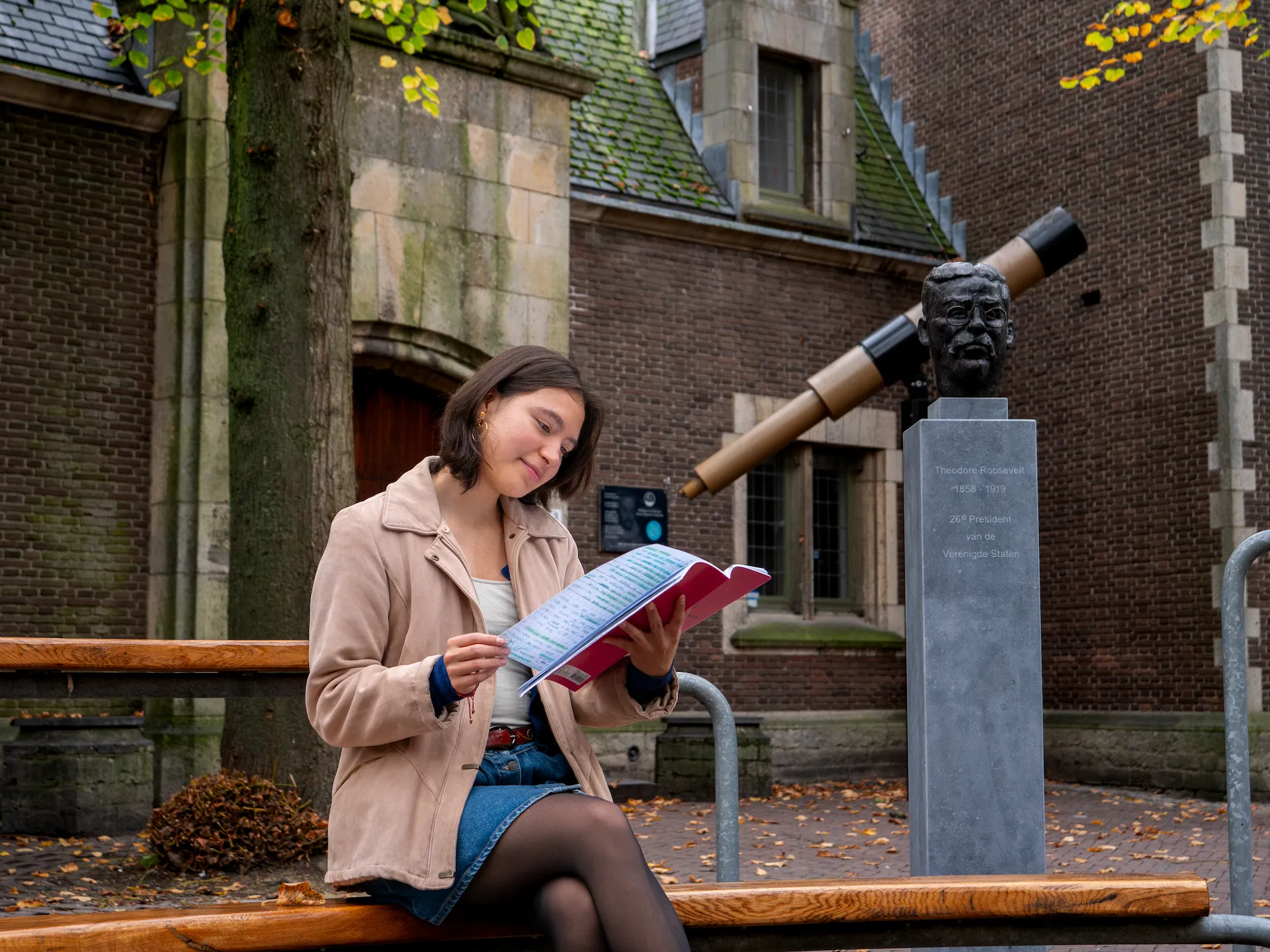 A student sitting at in the outdoor classroom of University College Roosevelt with a statue of Theadore Roosevelt behind her